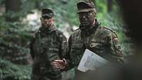 Ein Soldat mit Tarnfarbe erteilt im Wald einen Befehl. Unter seinem linken Arm hält er eine Karte.