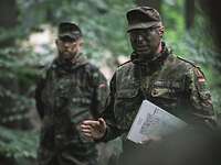 Ein Soldat mit Tarnfarbe erteilt im Wald einen Befehl. Unter seinem linken Arm hält er eine Karte.