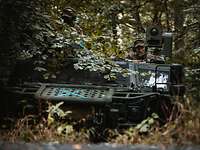 Im Wald steht gut versteckt ein Spähwagen, aus der Kommandantenluke schaut ein Soldat.