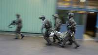 Vier Soldaten tragen einen Verletzten in einem Bergetuch an einer Lagerhalle vorbei.