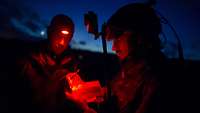 Ein Soldat hält unter Rotlicht Zettel und Stift in der Hand, auch ein zweiter trägt eine Stirnlampe.