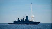 Ein graues Kriegsschiff in See, aus seinem Vorschiff ist eine Rakete gestartet, die eine Rauchspur hinter sich her zieht.