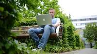 Ein junger Mann sitzt in seiner Freizeit draußen auf einer Bank vor einem Laptop.