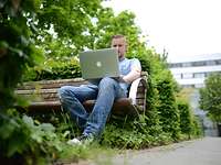Freizeit- und Kulturangebot Ein junger Mann sitzt in seiner Freizeit draußen auf einer Bank vor einem Laptop.