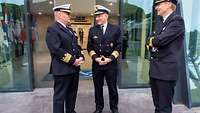 Drei Offiziere in dunkelblauen Marineuniformen und weißen Schirmmützen stehen im Eingangsbereich eines Bürogebäudes.
