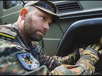 Ein Soldat hockt vor einem Reifen eines Geländewagen und hört einem zweiten Soldaten zu. 