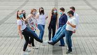 Eine Gruppe junger Menschen mit Mund-Nasenschutz-Bedeckungen in einem Kreis stehend und sich mit jeweils einem Fuß berührend