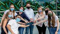 Im Halbkreis stehende Gruppe Menschen mit Mund-Nasenschutz-Bedeckungen strecken jeweils einen Arm in die Mitte aus