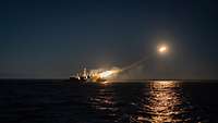 Auf dunkler See feuert ein Schiff eine rötlich beziehungsweise orange leuchtende Rakte ab.