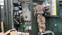 Ein Soldat steht auf einem Arbeitspodest und hantiert an Armaturen hinter einer geöffneten Wartungsklappe