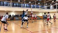 Volleyballer spielen in einer Halle. Im Hintergrund sitzen Zuschauer auf Bänken.