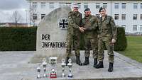 Drei Soldaten reichen sich die Hände. Vor ihnen Pokale, hinter ihnen der Stein der Infanterie.