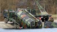 Ein Brückentransporter steht mit aufgefahrenem Ladebaum am Ufer, um einen Innenabschnitt der Faltschwimmbrücke aufzunehmen.