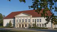 Ein beiges Gebäude steht im Sonnenschein, davor Rasen und ein Baum.