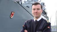 Ein Mann steht in blau-weißer Marineuniform vor einem grauen Schiff. 