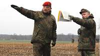 Zwei Soldaten stehen auf einem freien Feld und weisen beide mit jeweils einem Arm in eine Richtung.
