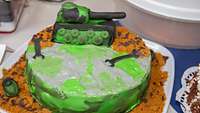 Eine Motivtorte mit einem grün-grauen Panzer und Werkzeugen aus Fondant steht auf einem Tisch.