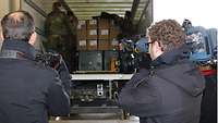 Menschen mit Kameras blicken in beladenen LKW, in dem eine Soldatin und ein Soldat Material für die Entladung vorbereiten.