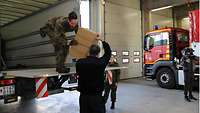 Soldat reicht einen Stapel Pakete von der Ladefläche eines LKWs an eine zivile Person.
