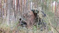 Ein Soldat liegt getarnt im Wald mit der Waffe im Anschlag