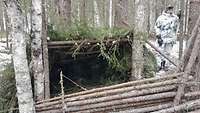 Zwischen Bäumen sind im Quadrat Holzstämme aufgestapelt. Als Windschutz dient eine Schräge aus Ästen