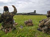 Zwei Soldaten sitzen mit ihrer Ausrüstung auf einer Wiese, ein Soldat hält den Daumen hoch, im Hintergrund ein Transportflugzeug