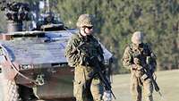 Zwei Soldaten laufen vor einem gepanzerten Fahrzeug und senken den Blick prüfend auf den Boden.