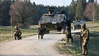 Vier Soldaten knien auf einem Weg und sichern ihre Sichtbereiche, hinter ihnen Gefechtsfahrzeuge.
