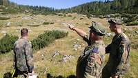 Drei Soldaten stehen auf dem Übungsplatz. Der mittlere Soldat zeigt ins Gelände.