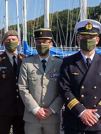 Vier Soldaten unterschiedlicher Nationen stehen am Bootshafen mit Mundschutzmasken vor Segelbooten und blicken in die Kamera.