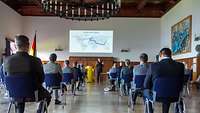 Die Lehrgangsteilnehmenden sitzen mit Abstand in der Aula. Flottillenadmiral Wilhelm Abry spricht über die Deutsche Marine.