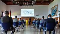 Die Lehrgangsteilnehmenden sitzen mit Abstand in der Aula. Flottillenadmiral Wilhelm Abry spricht über die Deutsche Marine.