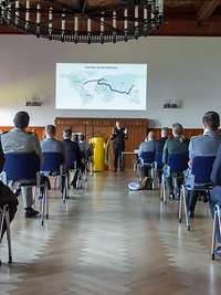 Die Lehrgangsteilnehmenden sitzen mit Abstand in der Aula. Flottillenadmiral Wilhelm Abry spricht über die Deutsche Marine.