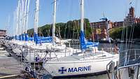 Im Bootshafen der Marineschule Mürwik liegen mehrere Segelboote. Im Hintergrund ist die Marineschule zu sehen. 