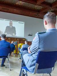 Die Lehrgangsteilnehmenden sitzen in der Aula und hören dem Stellvertreter des Inspekteurs der Marine per Videokonferenz zu.