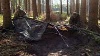 Zwei Soldaten bauen im Wald mit Zeltstangen und einer Zeltplane ein Versteck.
