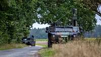 Ein Militärfahrzeug mit hoch aufragender Optronik steht am Rand einer Feldstraße, ein zweites folgt.