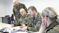 Eine Gruppe von Soldaten sitzt und steht an einem Tisch voller Arbeitspapiere und diskutiert.