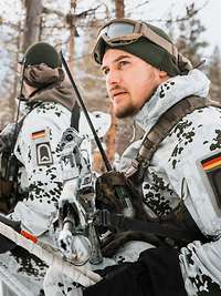 Kampf im Winter Zwei Soldaten stehen in Schneetarnanzügen, mit Schneestöcken und Waffen am Rand eines Waldes.