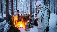 Mehrere Personen wärmen sich an einer Feuerstelle im verschneiten Wald.