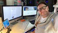  Der Redakteur, ein Mann mit Bart, schwarzer Brille und Cap, an seinem Homeoffice-Arbeitsplatz.