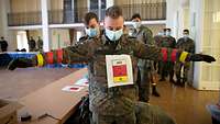 Soldaten mit Atemschutzmasken in einem Raum. Ein Soldat breitet die Arme aus und trägt rote und gelbe Bänder an den Armen