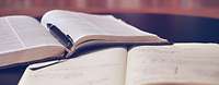 Zu sehen sind zwei aufgeschlagene Bücher, die übereinander liegen. Im Hintergrund ist ein Bücherregal zu erkennen.