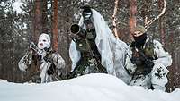 Soldaten hocken getarnt mit einer Panzerabwehrrakete und Handwaffen auf einem schneebedeckten Hügel.