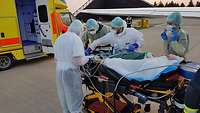 Vier Personen in Schutzanzügen mit Masken stehen um einen Patienten auf einer Transporttrage 