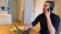 Ein Mann mit Telefon am Ohr, an einem Tisch sitzend.