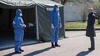 Zwei Menschen in blauer Schutzkleidung mit Mundschutz stehen vor einem Zelt einem Mann gegenüber.