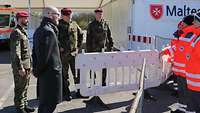 Soldaten, ein Zivilist und drei Menschen in Warnjacken stehen beieinander, dazwischen Absperrgitter.