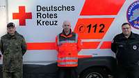 Drei Männer stehen vor einem weiß-orangefarbenen Rettungsfahrzeug des DRK.