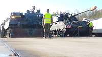 Ein Bergepanzer und eine Panzerhaubitze nebeneinander an der Entladerampe, davor ein Entladehelfer.